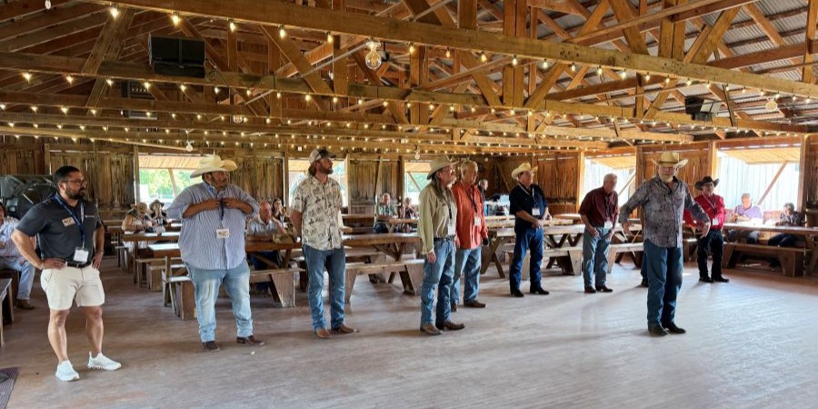 Photo shows a line of men standing on a dance floor. Behind them are several tables and benches.