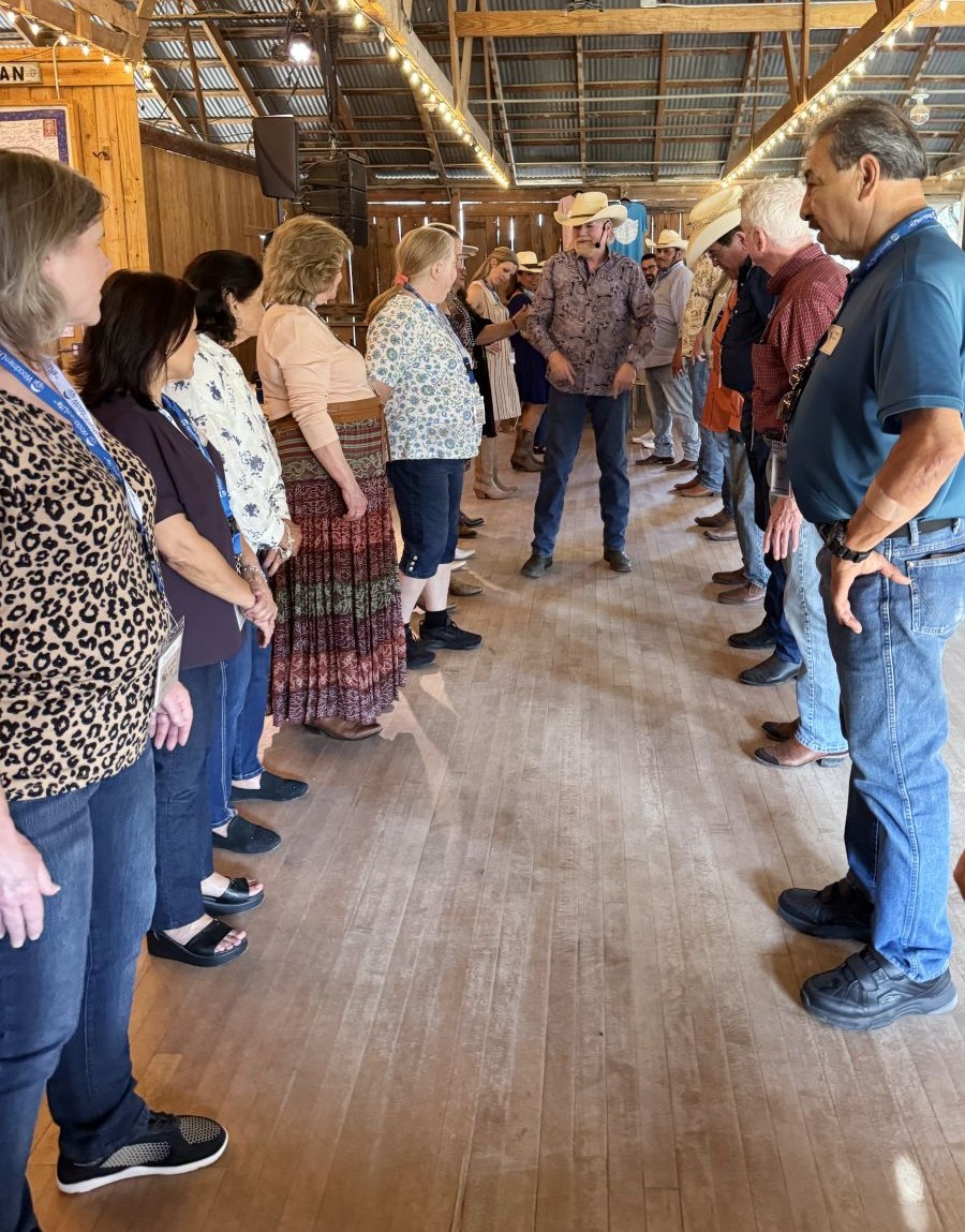 Photo is looking down the middle of two rows of people. Women are lined up on the left, and men are lined up on the right. The lines of people are facing each other. At the far end of the two lines stands a man wearing a cowboy hat. People are facing him for instructions on the dance.