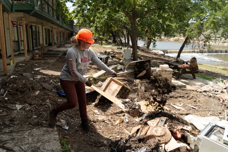 Photo shows a woman shoveling debris left behind after a natural disaster. She is wearing a grey "Team Rubicon" shirt and an orange hardhat. Behind her is a waterway and trees.