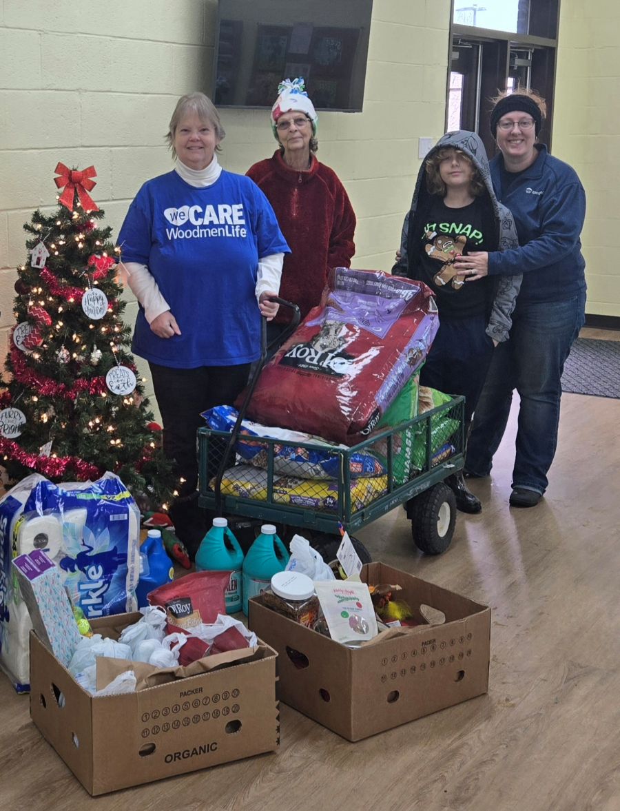 Photo shows four people — three adults and one boy — standing next to a small Christmas tree and boxes full of donations.