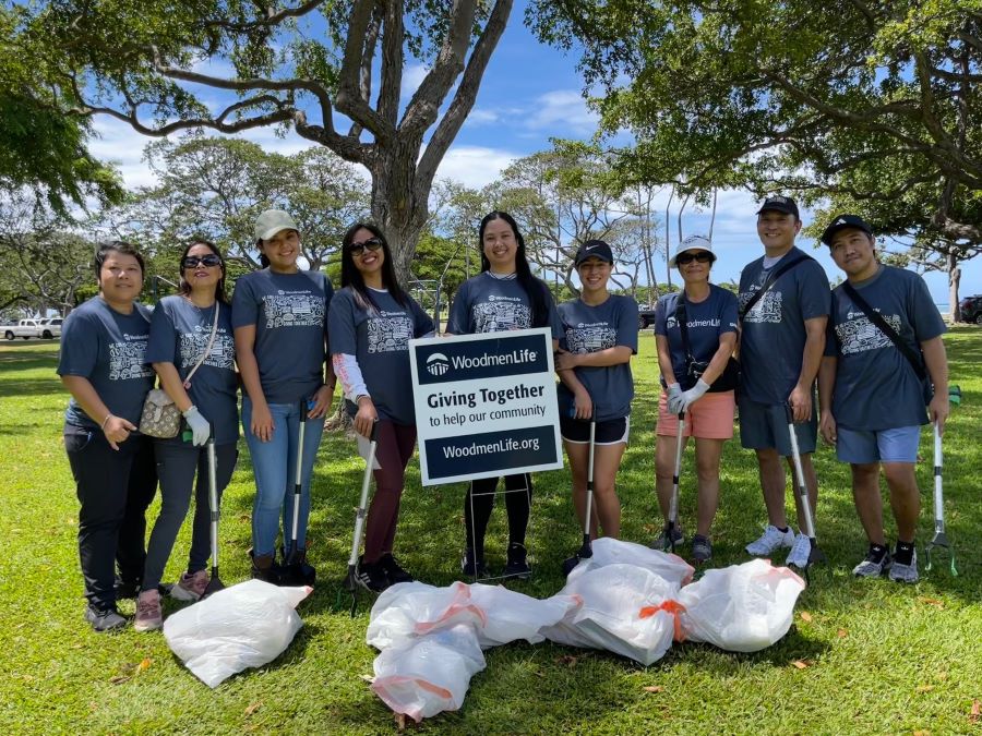 Nine people stand together with a sign that says "WoodmenLife. Giving Together to help our community. WoodmenLife.org." There are several filled trash bags in front of them.