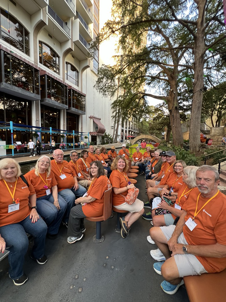 Photo shows about 20 people sitting along the sides of a long boat. They are all wearing orange T-shirts.