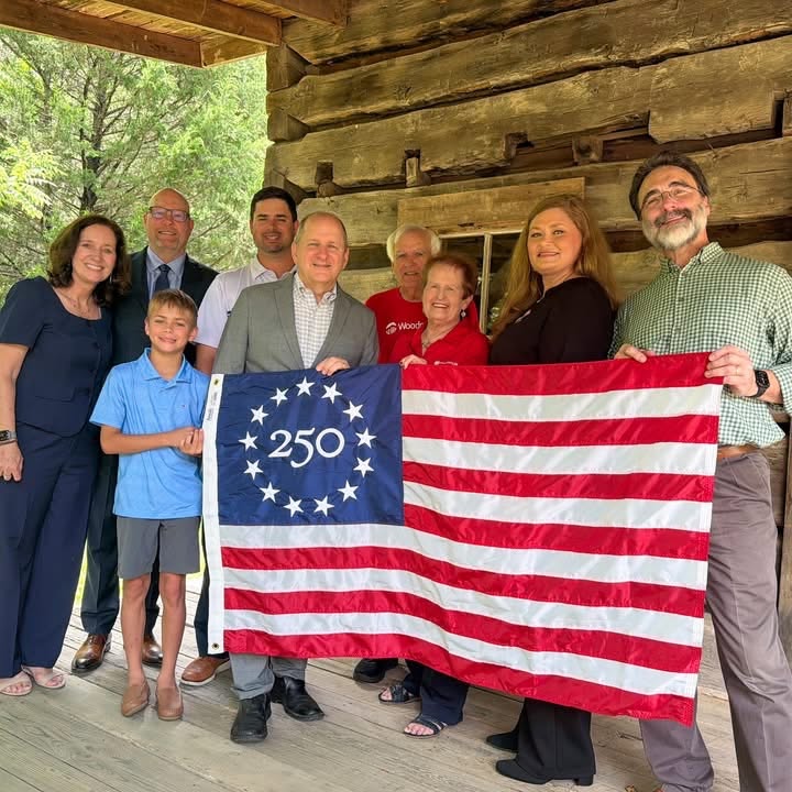 A group of nine people stand together holding a commemorative Betsy Ross 250 U.S. flag. They are standing in front of a wood-paneled building and there are trees in the background.