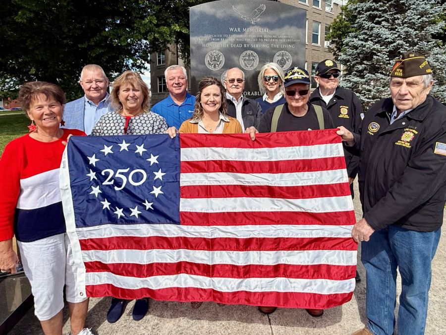 A group of 10 people stand together holding a commemorative Betsy Ross 250 U.S. flag. The group is standing outside in front of a war memorial.