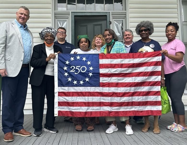 A group of nine people stand together holding a commemorative Betsy Ross 250 U.S. flag. They are standing on a porch, with a building and door behind them.
