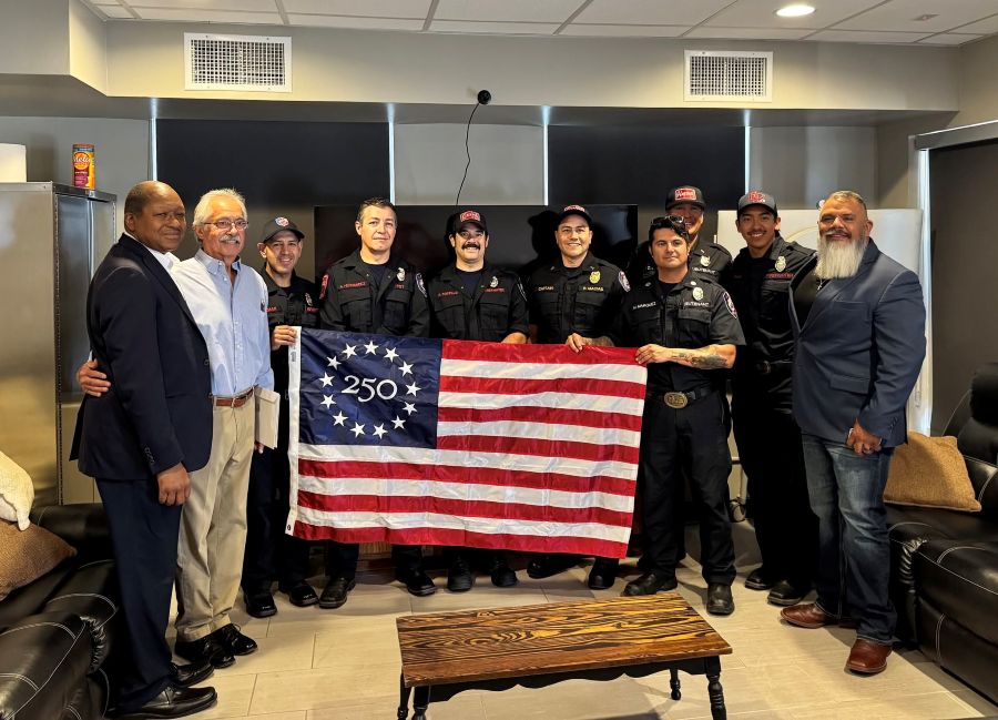 A group of 10 people stand together with a commemorative Betsy Ross 250 U.S. flag. Seven of the people are wearing police uniforms.