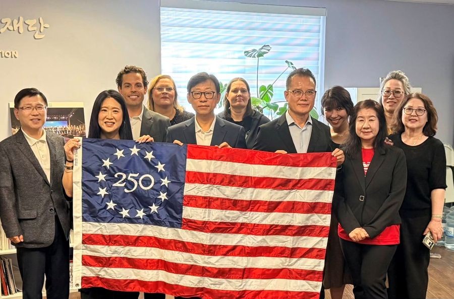 A group of 11 people stand together with a commemorative Betsy Ross 250 U.S. flag. They are standing inside, and there is a window behind them.