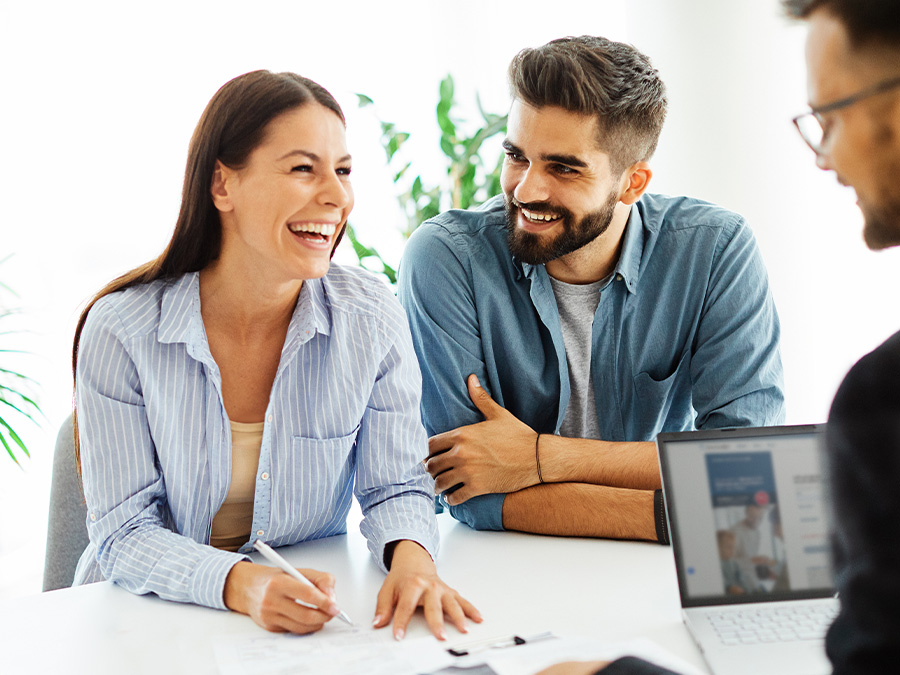 Stock photo shows a man and a woman sitting on one side of a table. The woman is holding a pen and writing on a piece of paper in front of her. Across from them is a man in a suit with a laptop open in front of him. All three people are smiling.