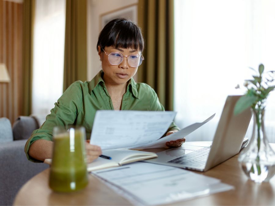 Stock photo shows a woman sitting at a table in a home. She is holding pieces of paper and looking over them. There is a laptop open on the table in front of her. Also on the table are a cup and a vase with a plant.