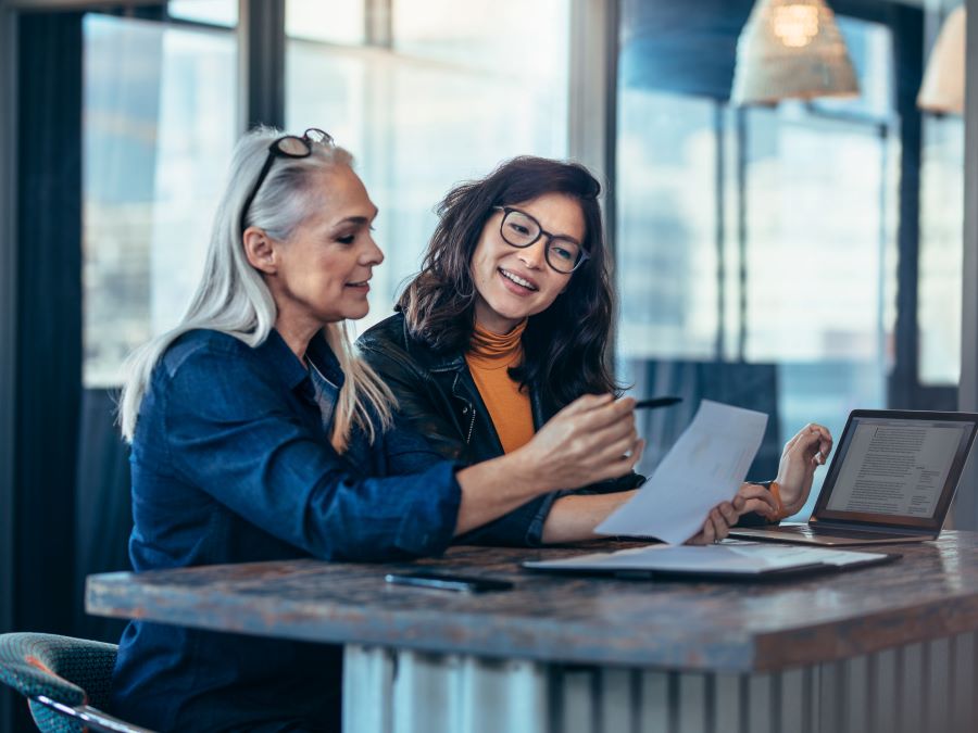 Stock photo shows two women sitting together at a table. One is holding a piece of paper and pointing toward something on the paper with a pen. Both women are smiling.