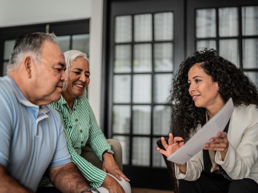 An older couple sit across from a woman. The woman is holding up a piece of paper for the couple to see and is pointing to something on the paper. Everyone looks happy and comfortable.