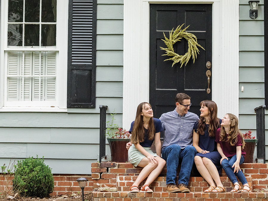 A family of four sits on the front steps of their home. A mother and father are in the middle, and a daughter sits on either side of them. Behind them is the front door to the home, which is adorned with a wreath.