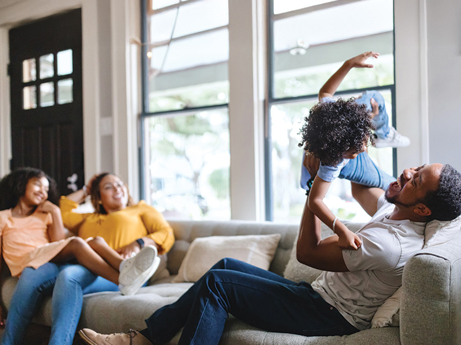 Stock photo shows a family sitting on a couch. On one end, the dad is lifting a child up in the air; on the other end, the mom is sitting with a daughter on her lap.