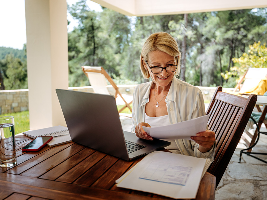 A woman sits at a table outdoors. On the table in front of her is an open laptop and papers she is looking at. Behind her are green trees. She is smiling.
