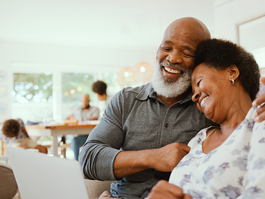 Stock photo shows an older man and woman sitting together. The man has his arm around the woman, and she is leaning into him. Both are smiling. In the foreground, the woman is holding up a piece of paper in front of them. In the background, people are gathered at a table.
