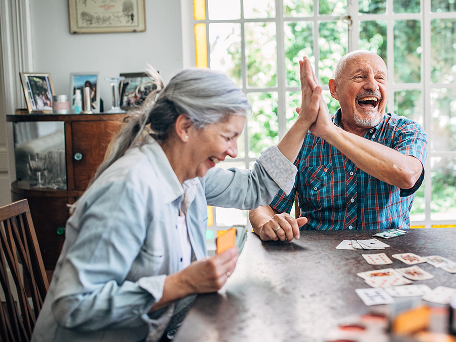 Stock photo shows two older people sitting at a table. They are laughing and high-fiving. Playing cards are laid out on the table in front of them.
