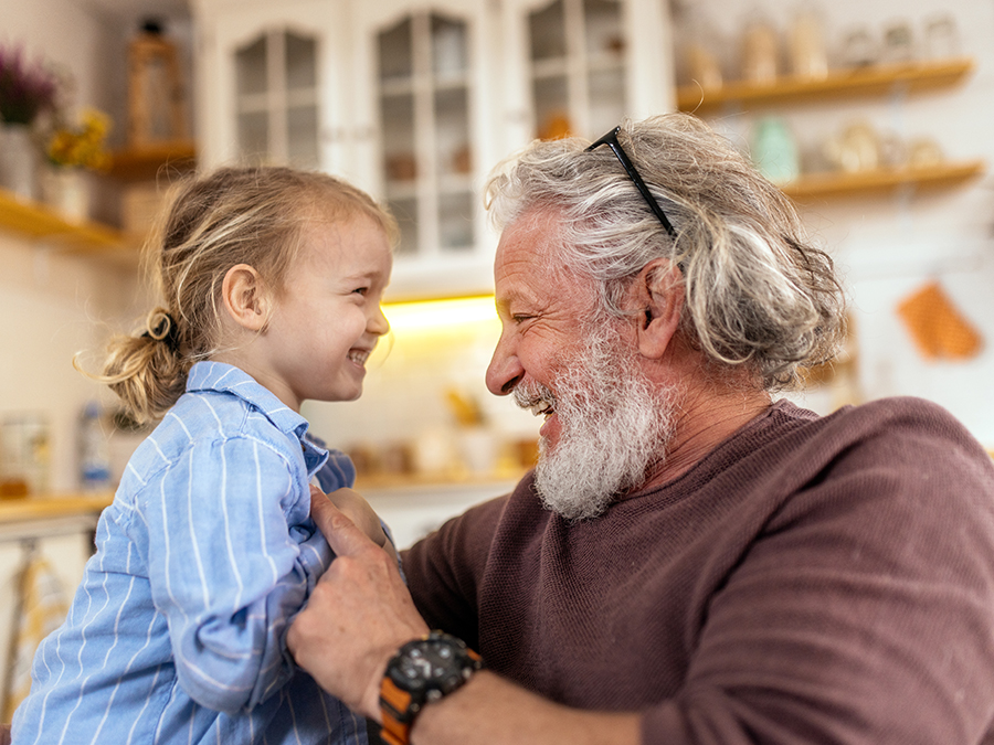 Stock photo shows an older man sitting with a young child. They are both smiling and laughing as the man pokes at the child. There are cabinets and shelves on the wall behind them.