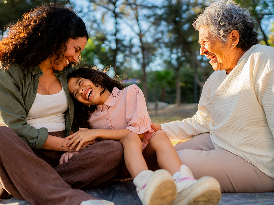 Stock photo shows three people: a middle-age woman, a young girl, and an older woman. They are sitting on a blanket outside. They are smiling and laughing together, and the young girl is leaning to the left, into the middle-age woman. Trees are visible behind the group.