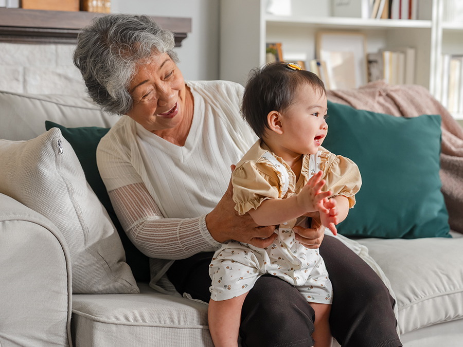 Stock photo shows an older woman sitting on a couch with an infant on her lap. The woman is smiling as she holds the child, and the child is smiling and clapping.
