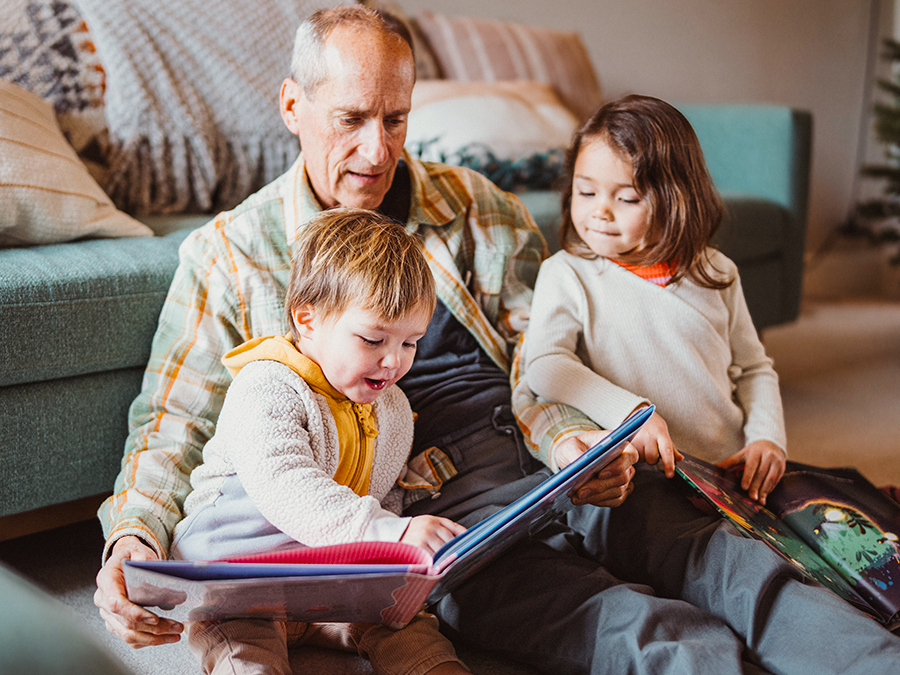 Stock photo shows an older man sitting on the ground with two children. They are sitting against a couch, and the man is holding open a picture book in front of them.
