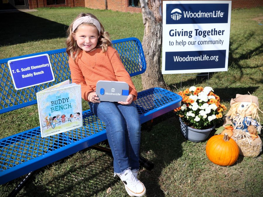 This picture shows an elementary school-aged girl sitting on a blue bench. There is a sign on the back of the bench that reads "T.G. Scott Elementary Buddy Bench." Sitting on the bench with the girl is a children's book called "The Buddy Bench." Behind the girl is a sign that says "WoodmenLife. Giving Together to help our community. WoodmenLife.org," as well as autumnal decorations like flowers and a pumpkin.