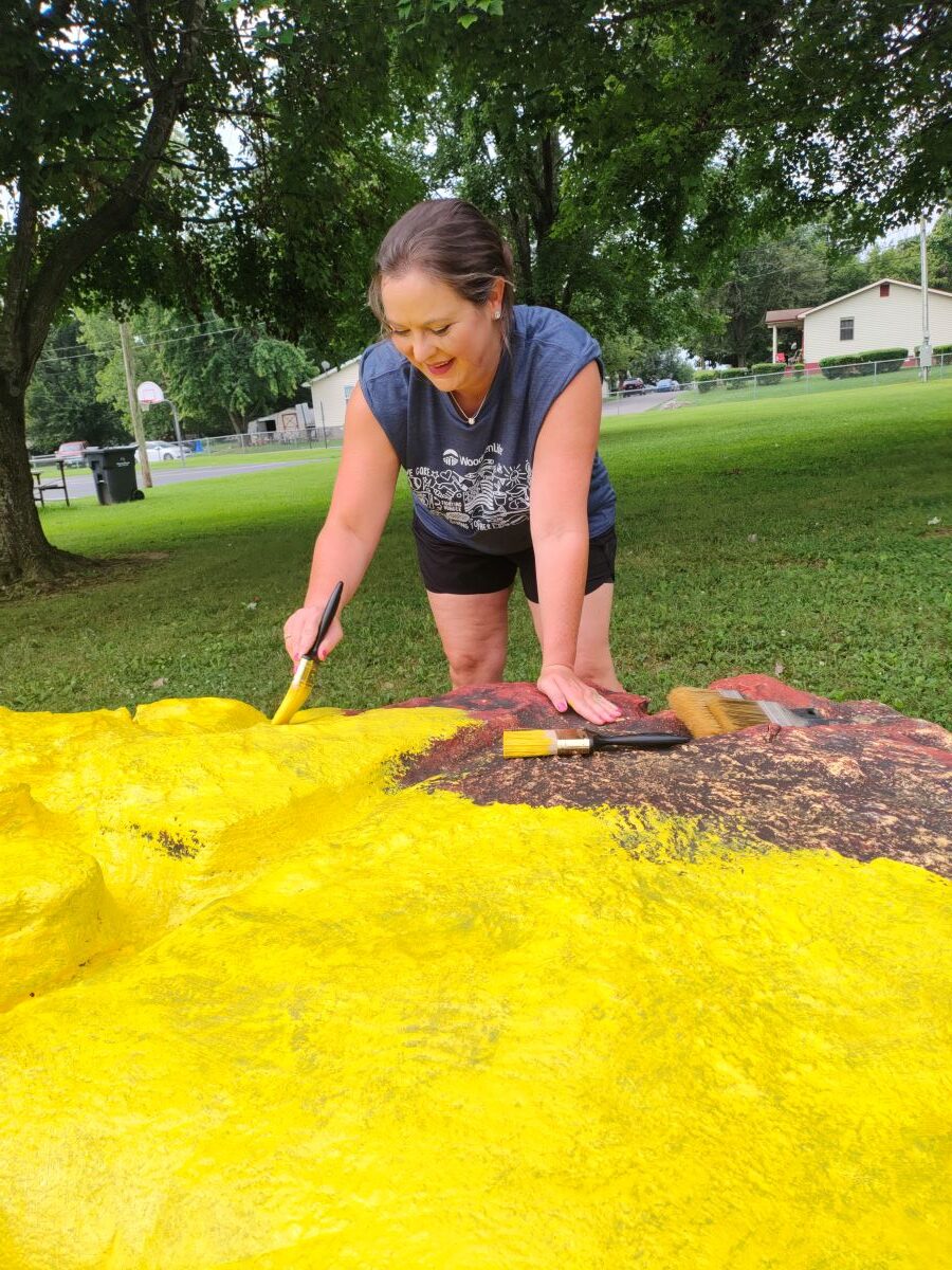 In this picture, a woman is painting a large rock yellow. She is wearing a blueish-gray WoodmenLife-branded t-shirt.