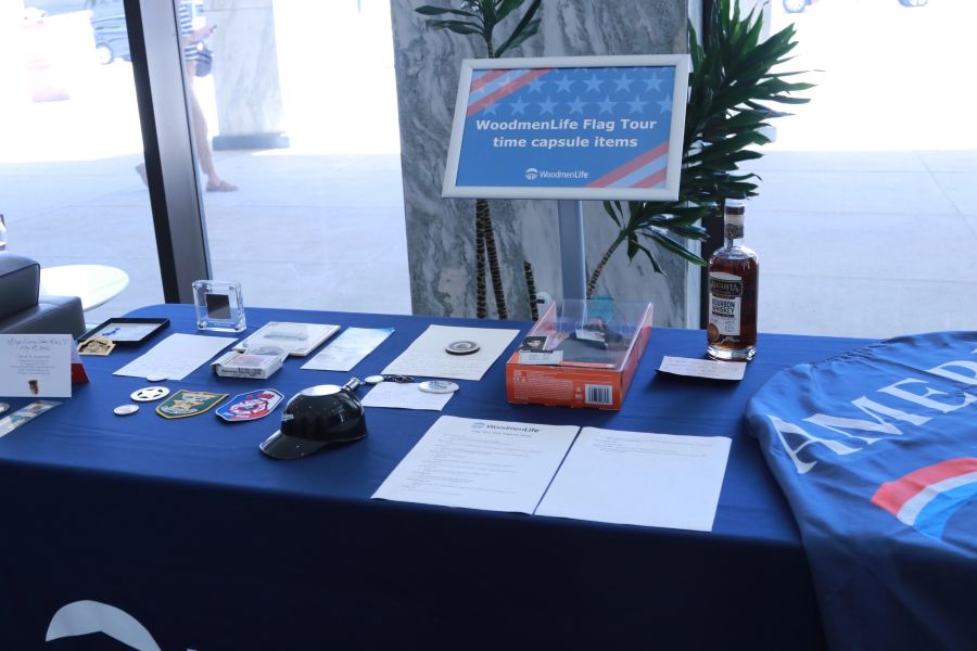 Photo shows a table covered in items collected for a time capsule. Included are patches, medals, a small plastic baseball helmet, a doll in a box, and a bottle of whiskey. Behind the table is a sign that says "WoodmenLife Flag Tour time capsule."