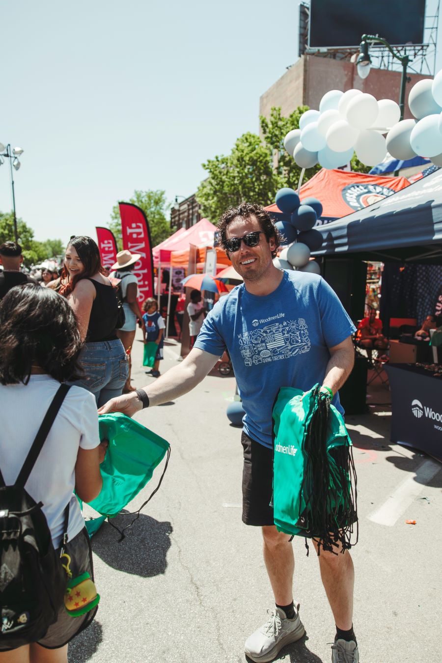 Photo shows a man in a blue WoodmenLife T-shirt handing out green drawstring backpacks to people walking by. Behind him are a row of tents/booths that people attending the Cinco de Mayo parade can visit. The sky is blue, and it appears bright and sunny out.
