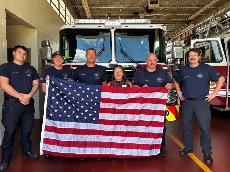 A WoodmenLife Representative stands with five firefighters. Together, they are holding an American flag. The group is standing in front of a fire truck.