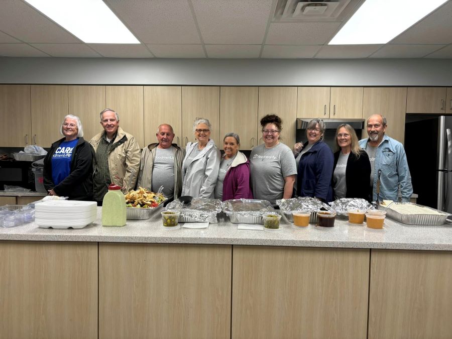 Nine people stand behind a counter. On the counter are large aluminum serving trays filled with food.