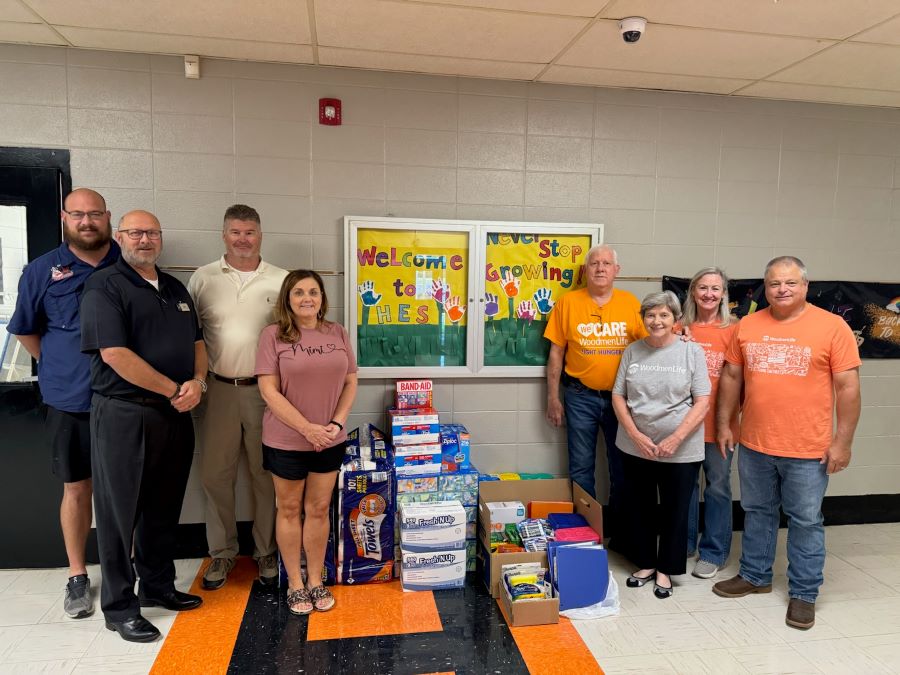 Eight people stand around a stack of donated school supplies, which includes paper towels, Band-Aids, binders, and markers. The group of people and school supplies are standing in an elementary school hallway.