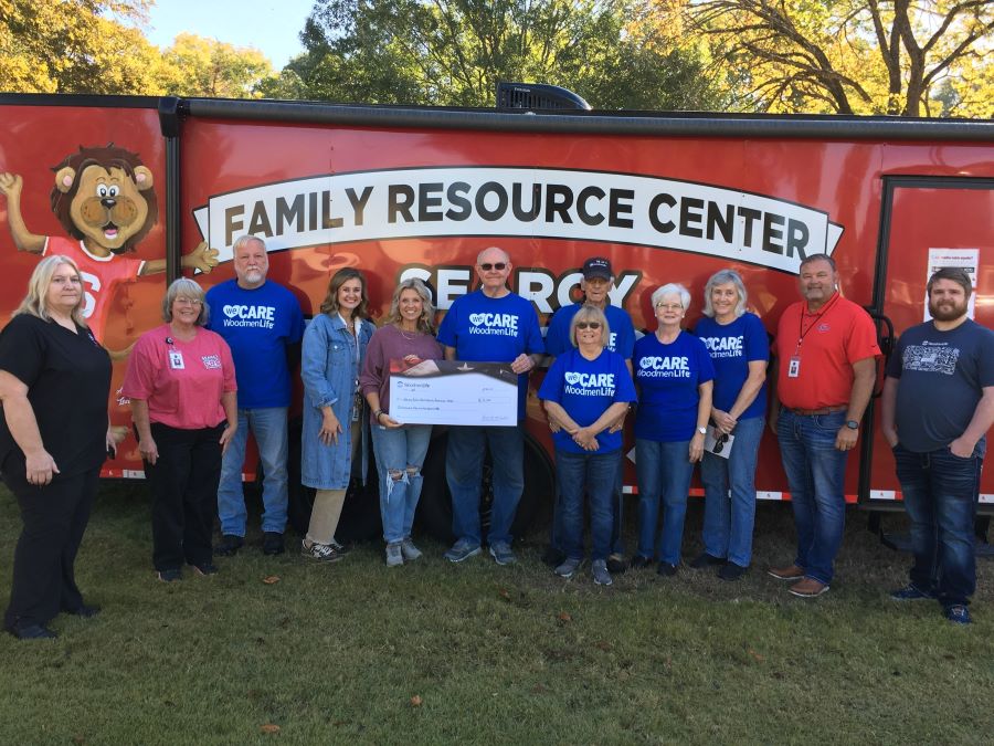 A group of 12 people stand in front of a red bus with the words "Family Resource Center. Two people in the middle are holding an oversized check. Several people are wearing blue t-shirts that say "WeCare WoodmenLife."