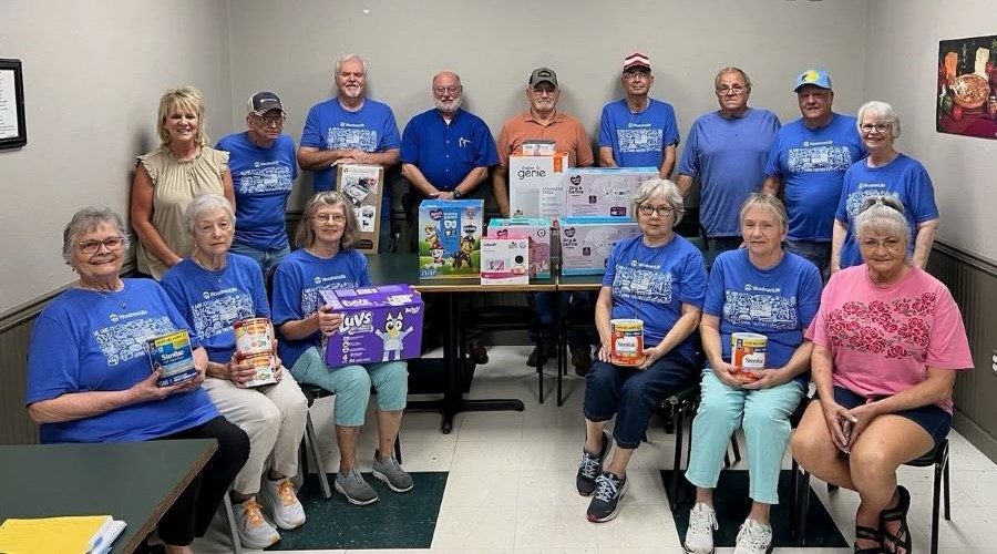 A group of 15 people, some standing and some sitting, gather around the donations. Several people wear blue WoodmenLife T-shirts.