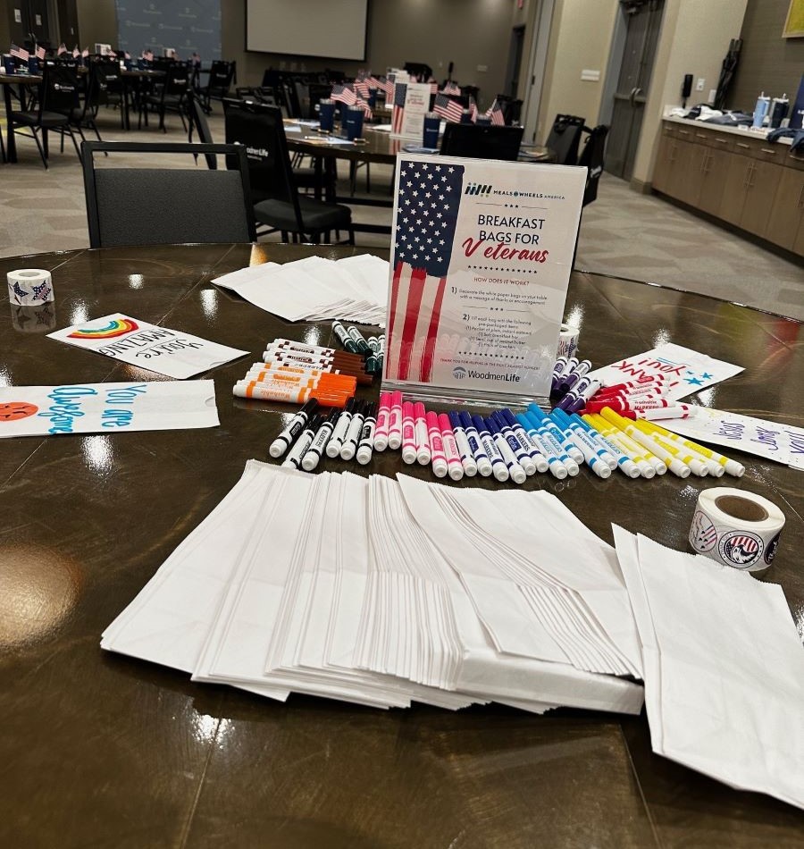 Photo shows a table on which is spread out plain white paper bags, lots of markers and rolls of stickers. In the middle of the table are instructions for the participants about decorating the bags.