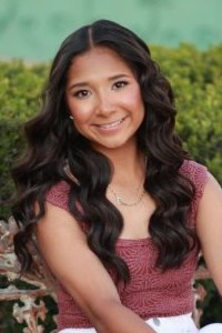 Photo is a senior portrait of Alazay M., a WoodmenLife member in Weimar, TX. She is wearing a deep red short-sleeved shirt, and there are green bushes behind her.