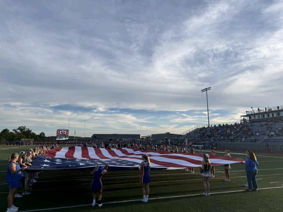 Photo show an oversized American flag being held open on display on a football field. People surround the flag, holding the edges. In the background, spectators in the bleachers look on.