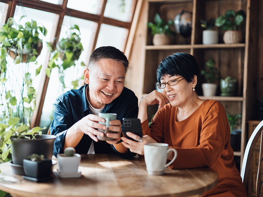 Stock photo shows an older couple sitting together at a table. He is holding a coffee mug, and she is holding a cellphone that they're both looking at. Behind them are several potted plants and a large window.
