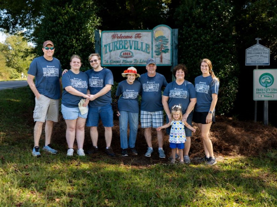 A group of eight people stand in front of a sign that says "Welcome to Turbeville." They are all wearing blue-gray WoodmenLife-branded t-shirts.