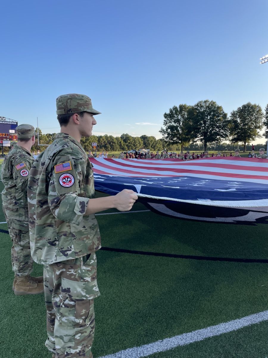 Photo shows a man in military fatigues holding up the edge of an oversized American flag. There are people alongside him and in the distance (across the flag) also holding it up. They are standing on an athletic field; there are trees and blue sky in the background.