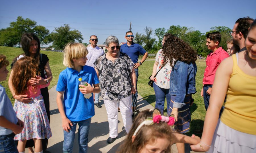 A woman is standing on a sidewalk, surrounded by her children and grandchildren. There is green grass on either side of the sidewalk, and trees in the background. The family members are greeting each other, hugging, and holding hands.