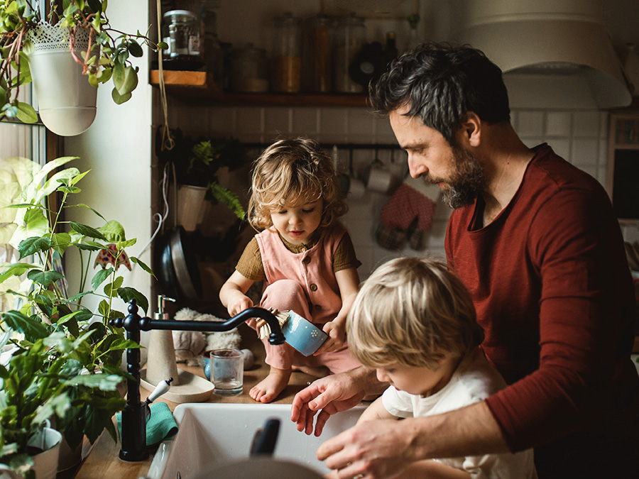 Stock photo shows a man and two young children at at kitchen sink. The man is facing the sink, with his hands reaching in; one child is between the man and the sink; the other child is sitting on the counter, facing the sink. There are plants on the window sill in front of them and kitchen gadgets on the wall behind them.
