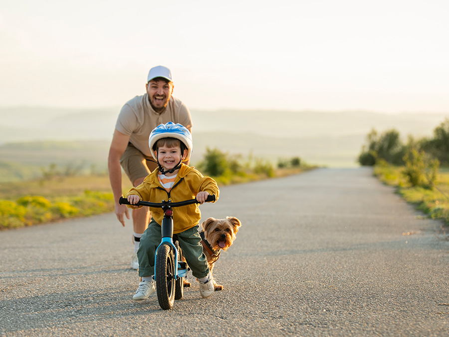 Stock photo shows an adult man standing behind and watching as a young boy rides a bicycle. There is a dog with them, and there is a long road behind them.
