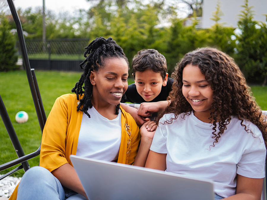 Stock photo shows a woman sitting outside with two children. A teenage girl sits beside the woman in a swinging chair, and a young boy stands behind the pair, looking over the back rest. All three are looking at a laptop on the teen girl's lap. Behind them are green grass and trees.