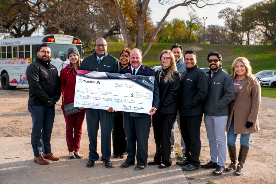 Photo shows 10 people standing together, with the middle two people holding an oversized check. The check is made out to Kroc Academy in the amount of $3,468. Behind them is a white school bus.
