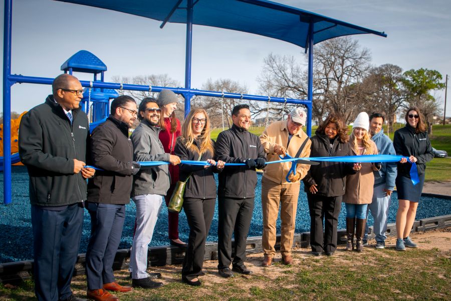 Photo shows 11 people standing in a row, holding a blue ribbon. A man in the middle is holding oversized scissors, about to cut the ribbon. Behind them is blue playground equipment.