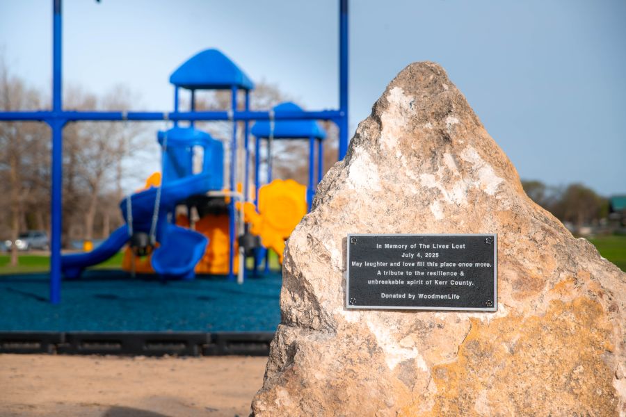 Photo shows a large rock with a plaque on it. The plaque reads: "In Memory of The Lives Lost, July 4, 2025. May laughter and love fill this place once more. A tribute to the resilience & unbreakable spirit of Kerr County. Donated by WoodmenLife." Behind the rock is a new playground.