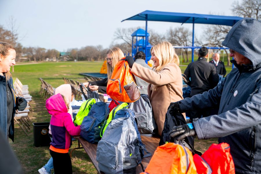 Photo shows a table filled with full backpacks. Adults on one side of the table are handing the backpacks to children on the other side of the table.