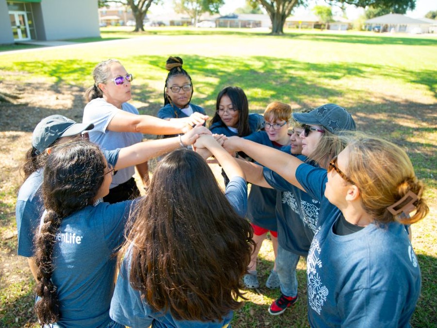 A group of people, both kids and adults, stand in a circle with their hands in.