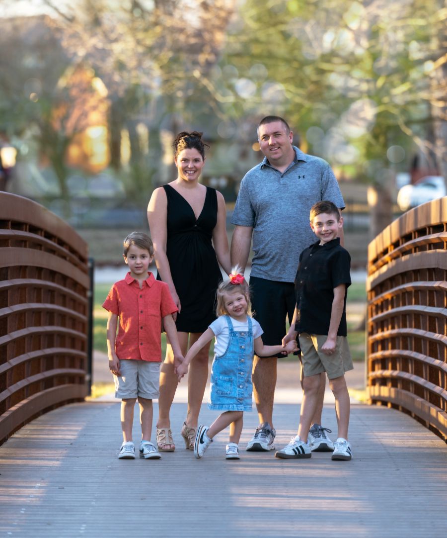 A family poses together on a bridge. The mom and dad stand behind their three children. Behind them are trees and other greenery.