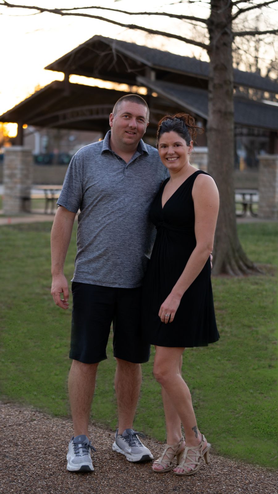 A man and a woman pose together for the photo. They are outside; behind them is green grass, a tree, and a park shelter.